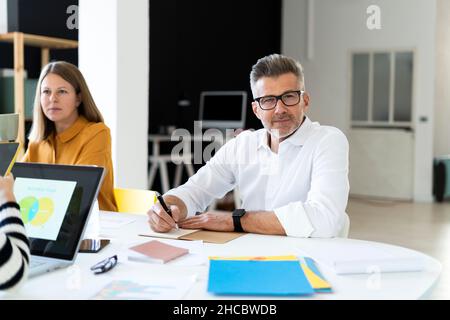 Thoughtful businessman with eyeglasses writing in diary sitting by colleague at office Stock Photo