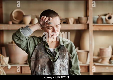 Female potter in apron looking very tired, she having a lot of work in pottery workshop Stock Photo