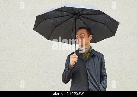 Horizontal medium shot of handsome Caucasian man wearing eyeglasses standing outdoors under umbrella looking away Stock Photo