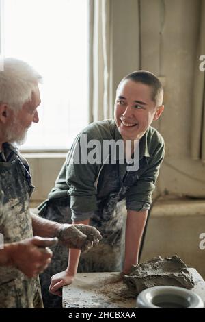 Young woman smiling to her colleague while he making sculpture from clay in workshop Stock Photo