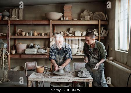 Senior man making earthenware dish on pottery wheel with woman giving him the piece of clay, they working in studio Stock Photo