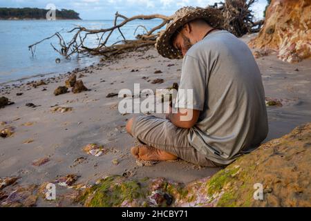Young white man in rustic hat sitting on the beach looking depressed. Stock Photo