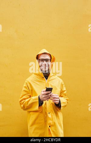 Vertical portrait of modern Caucasian man wearing yellow raincoat standing against yellow wall holding smartphone looking at camera Stock Photo