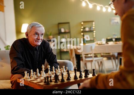 Pair of old Caucasian men siting in beautiful living room with served dining table on background and playing chess Stock Photo