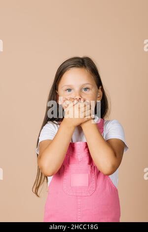 Portrait photo of little girl closing mouth with hands not talk with others with shining eyes looking at camera wearing bright pink jumpsuit and white Stock Photo
