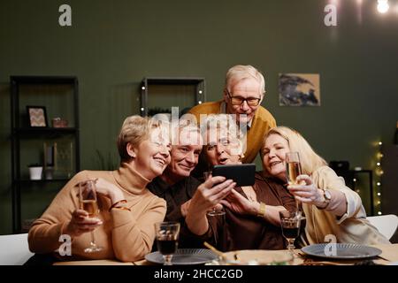 Group of five cheerful senior friends gathered at dining table for celebration, holding glasses and making selfie on smartphone camera Stock Photo