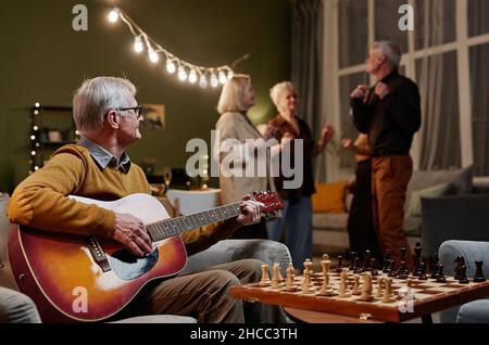 Old man sitting on armchair and playing acoustic guitar while his four friends dancing on background Stock Photo