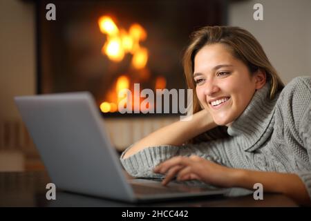 Happy woman using laptop watching media content at home near fireplace Stock Photo