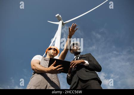 Indian engineer in uniform and helmet using tablet during meeting with african american inspector in stylish suit. Two partners discussing green energy while standing on farm with windmills. Stock Photo