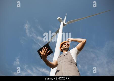 Indian engineer in orange helmet and grey overalls standing among windmill farm with modern tablet in hands. Foreman examining and controlling work of huge turbines. Stock Photo