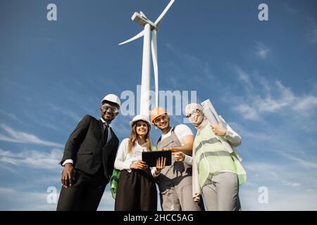 Group of four multiracial coworkers in safety helmets smiling on camera while staning on farm with huge windmills. Men and women using digital tablet and blueprints during meeting outdoors. Stock Photo