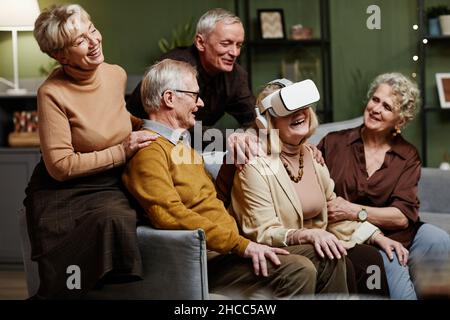 Senior Caucasian woman sitting on sofa and enjoying modern VR glasses while her four friends gathered around her and watching her Stock Photo