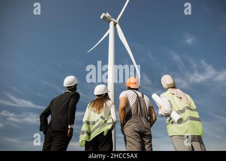 Back view of men and women in uniform and safety helmets looking on huge wind turbine while standing at rural area. Four multicultural partners discussing production of clean energy. Stock Photo