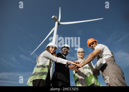 Group of four multicultural partners stacking hands together and smiling on camera while standing among wind turbines. Men and women in safety helmets having successful meeting outdoors. Stock Photo