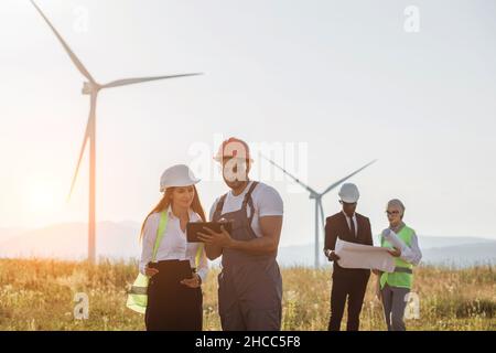 Team of multiracial industrial workers in safety helmets controlling process of green energy production on farm with wind turbines. Four colleagues using blueprints and tablet outdoors. Stock Photo