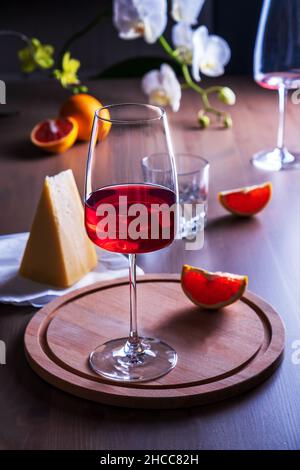 Elegant goblet with rose wine, cheese and oranges on wooden surface in romantic setting against backdrop of blooming orchids. Stock Photo
