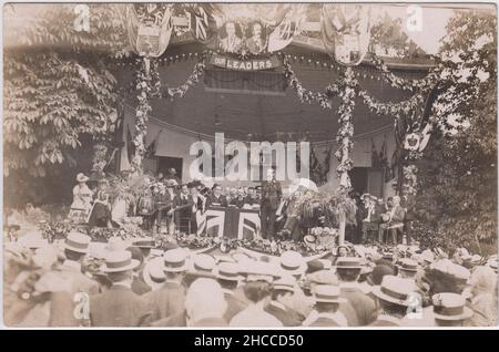 Bristol political meeting, early 20th century: a large crowd (many wearing boaters) are looking towards a platform with men and women seated, one man is standing up & speaking to the audience. The platform is decorated with Union Jack flags, garlands of flowers & foliage, coats of arms and portraits of 'our leaders', including Joseph Chamberlain. The photograph was taken by A.E. Smith of 44 Coronation Road, Bristol Stock Photo