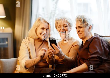 Group of three happy aged Caucasian women sitting together in living room against window and using smartphone Stock Photo