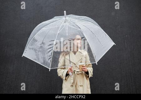 Horizontal medium shot of modern young woman wearing trench coat holding transparent umbrella and cup of coffee standing outdoors against dark gray wa Stock Photo
