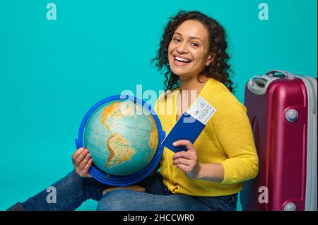 Cheerful woman traveler in yellow sweater sits next to luggage, holds globe, boarding pass, ticket and passport, smiles toothy smile looking at camera Stock Photo