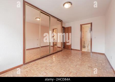 Empty bedroom with white walls with a small slatted parquet floor, a large built-in wardrobe with sliding mirrored doors and exit to the side of the b Stock Photo