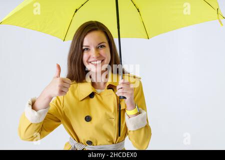 Joyful young woman holding umbrella and giving thumbs up Stock Photo