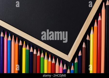 Close up of many wooden crayons in different colors lying under a dark blackboard with a wooden frame creating an outline with space for text