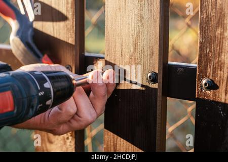 Hand of carpenter holding stud driving machine putting screw into wooden plank to metal construction for reconstructing wooden fence. Outdoor works. C Stock Photo
