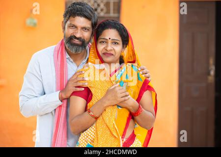 Portrait of young happy indian rural farmer couple standing outside house. Smiling village husband wife in traditional cloths looking at camera, beard Stock Photo