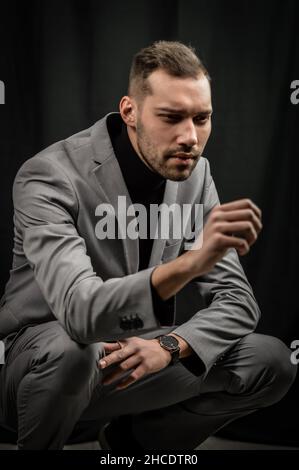 portrait of a young man in a gray suit and a black turtleneck, crouched on the ground looking intently Stock Photo