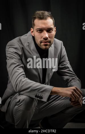 portrait of a young man in a gray suit and a black turtleneck, crouched on the ground looking intently Stock Photo