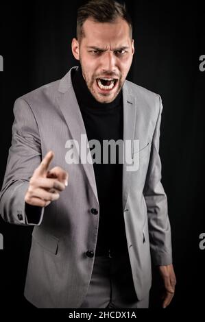 portrait of a young businessman in a gray suit, very angry yelling and gesturing accusingly in front of him Stock Photo