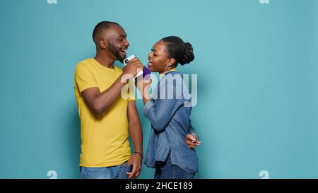 Playful couple singing song and using smartphone as microphone. Man and woman enjoying music and technology to have fun with mobile phone and dancing on sound, standing over blue background. Stock Photo
