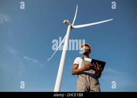 Competent technician using digital tablet while controlling working process of wind turbines outdoors. African american man wearing grey overalls, orange helmet and safety glasses. Stock Photo