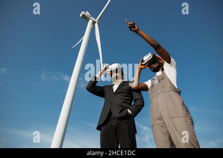 Indian businessman in suit and african american engineer in uniform using VR goggles for work on farm with wind turbines. Production of green energy and modern technology concept. Stock Photo