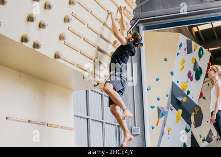 Full body side view of sportive barefoot male in activewear practicing rock climbing on wall with wooden grips in light gym Stock Photo