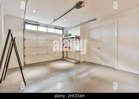 Spacious modern light garage with white closed automatic gate and special equipment placed on metal shelves near doors with minimalistic interior Stock Photo