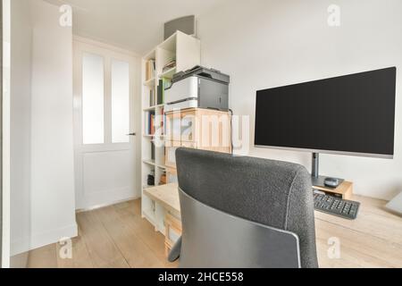 Comfortable chair placed at wooden desk with modern computer near bookshelf in workroom in daylight Stock Photo