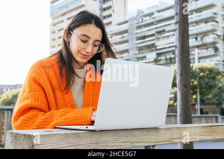 Young brunette female in bright cardigan orange typing on laptop keyboard while working remotely in terrace Stock Photo
