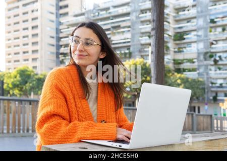 Young brunette female in bright cardigan orange typing on laptop keyboard while working remotely in terrace Stock Photo