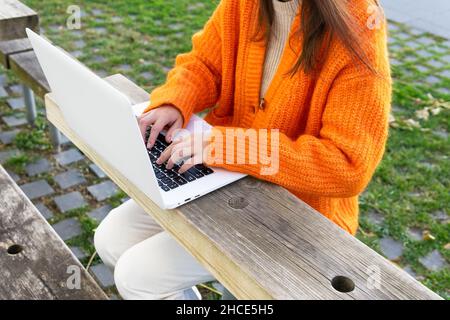 Crop unrecognizable female in bright cardigan orange typing on laptop keyboard while working remotely in terrace Stock Photo