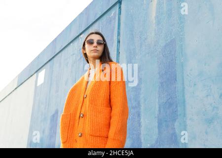 From below young female in fashionable sunglasses and bright orange knitted cardigan standing near blue wall Stock Photo