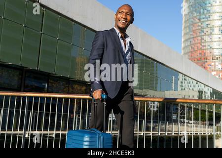 Side view of serious African American male entrepreneur in formal suit strolling on footpath with luggage near lake in city Stock Photo