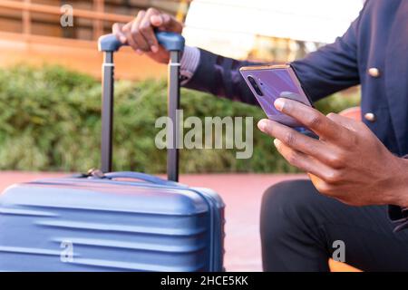 Cropped unrecognizable African American male entrepreneur with luggage wearing elegant wear surfing modern cellphone on street with modern building in Stock Photo