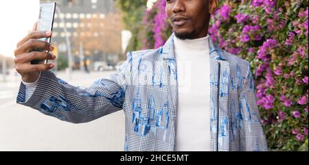 Cropped unrecognizable African American man in stylish outfit talking self portrait on modern smartphone while standing near blooming bushes on street Stock Photo