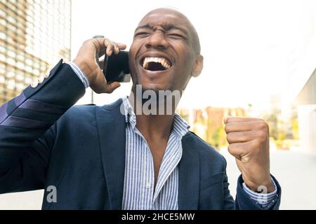 Joyful African American male entrepreneur in suit having phone conversation at table with coffee on terrace of cafe Stock Photo