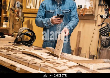 Crop faceless male carpenter in denim uniform using mobile phone while working in professional workshop Stock Photo