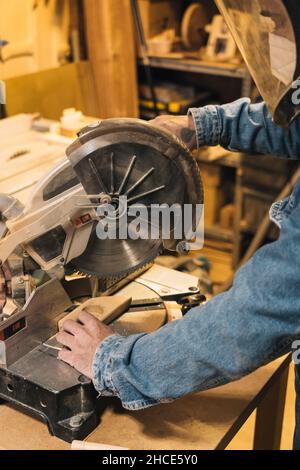 Side view of crop anonymous male carpenter in protective mask and denim overall cutting wood with miter saw in workshop Stock Photo
