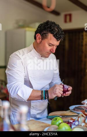 Concentrated adult male chef in white uniform cutting onion while cooking and standing near table with ingredients in light kitchen Stock Photo