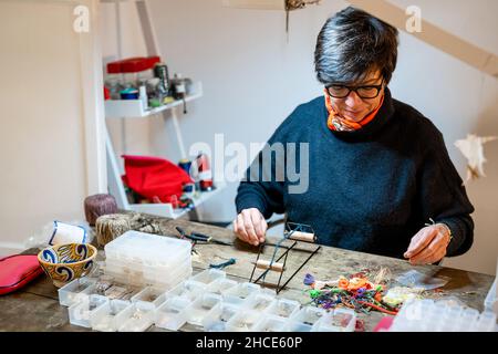 Positive Woman using professional instrument with leather cord while making bijouterie at table with various decorative elements in workshop Stock Photo